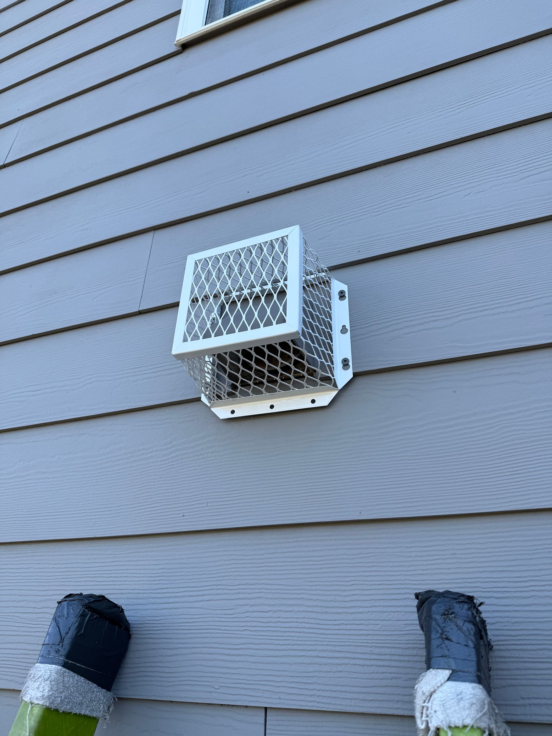 Wall-mounted outdoor metal vent on gray house siding above two wrapped pipe openings