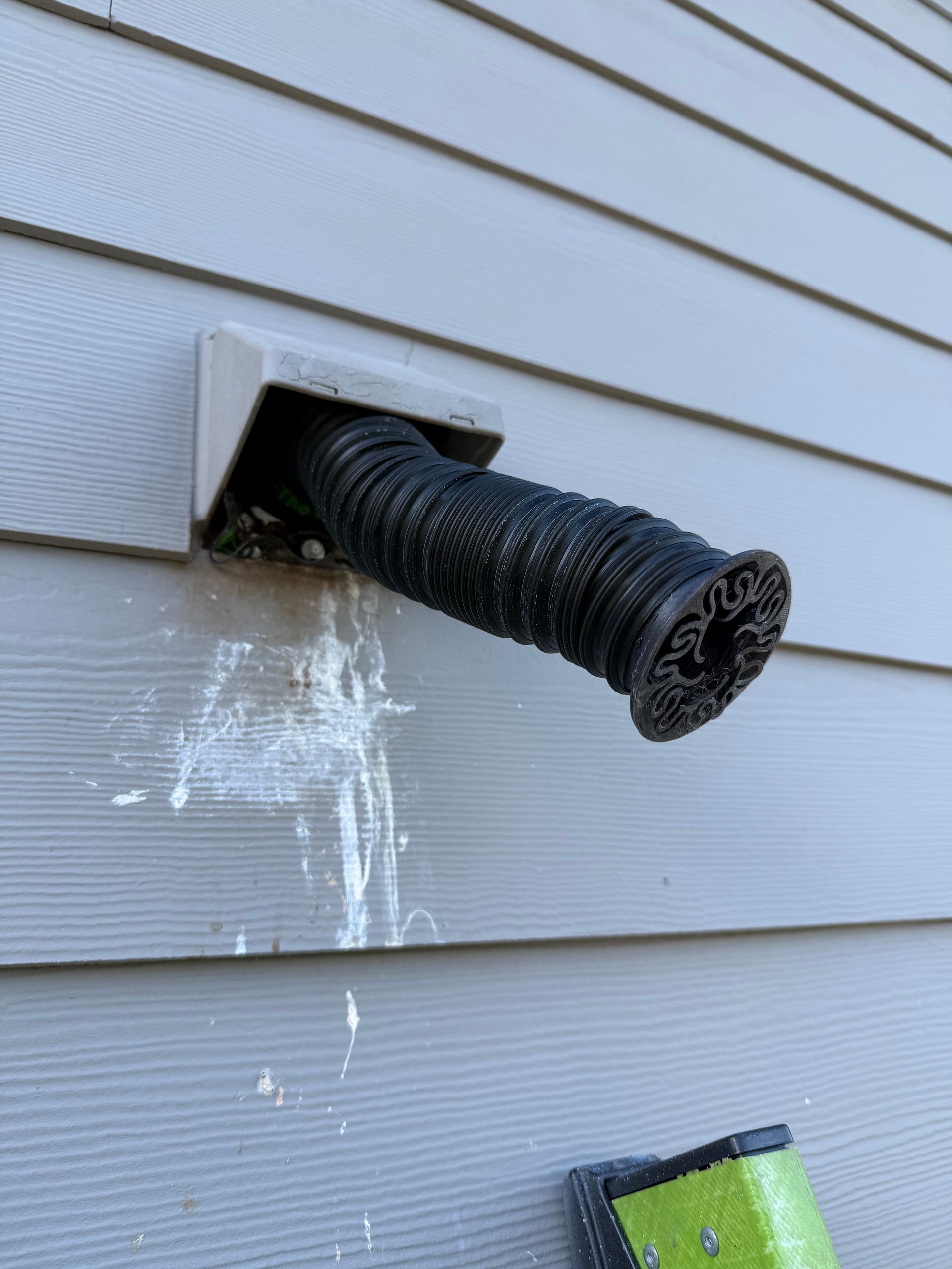 Black flexible vent pipe protruding from an exterior wall with white siding and water stains below.