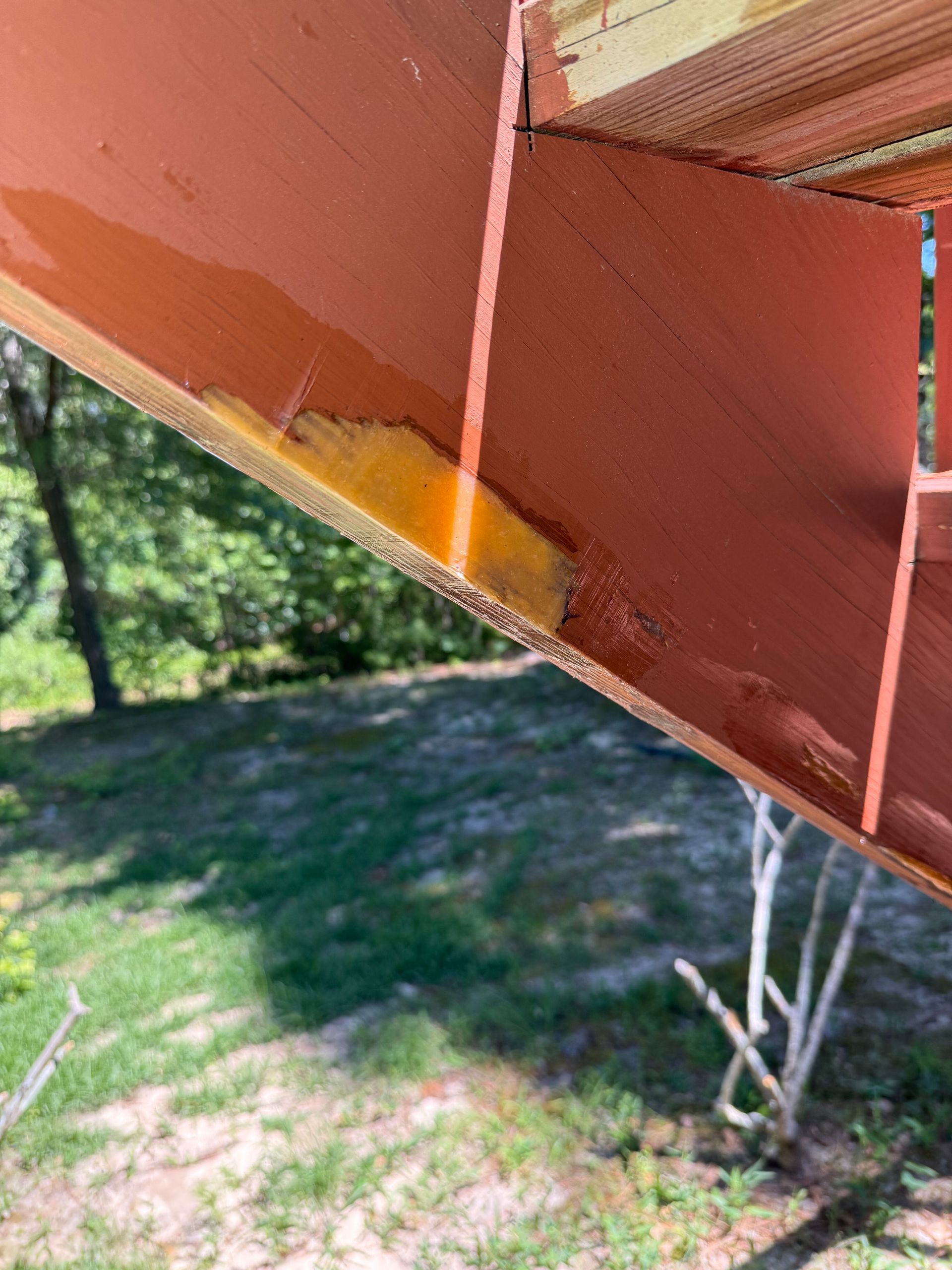 Close-up of a rust-red metal roof or beam with a yellow paint streak, viewed against trees and grass below