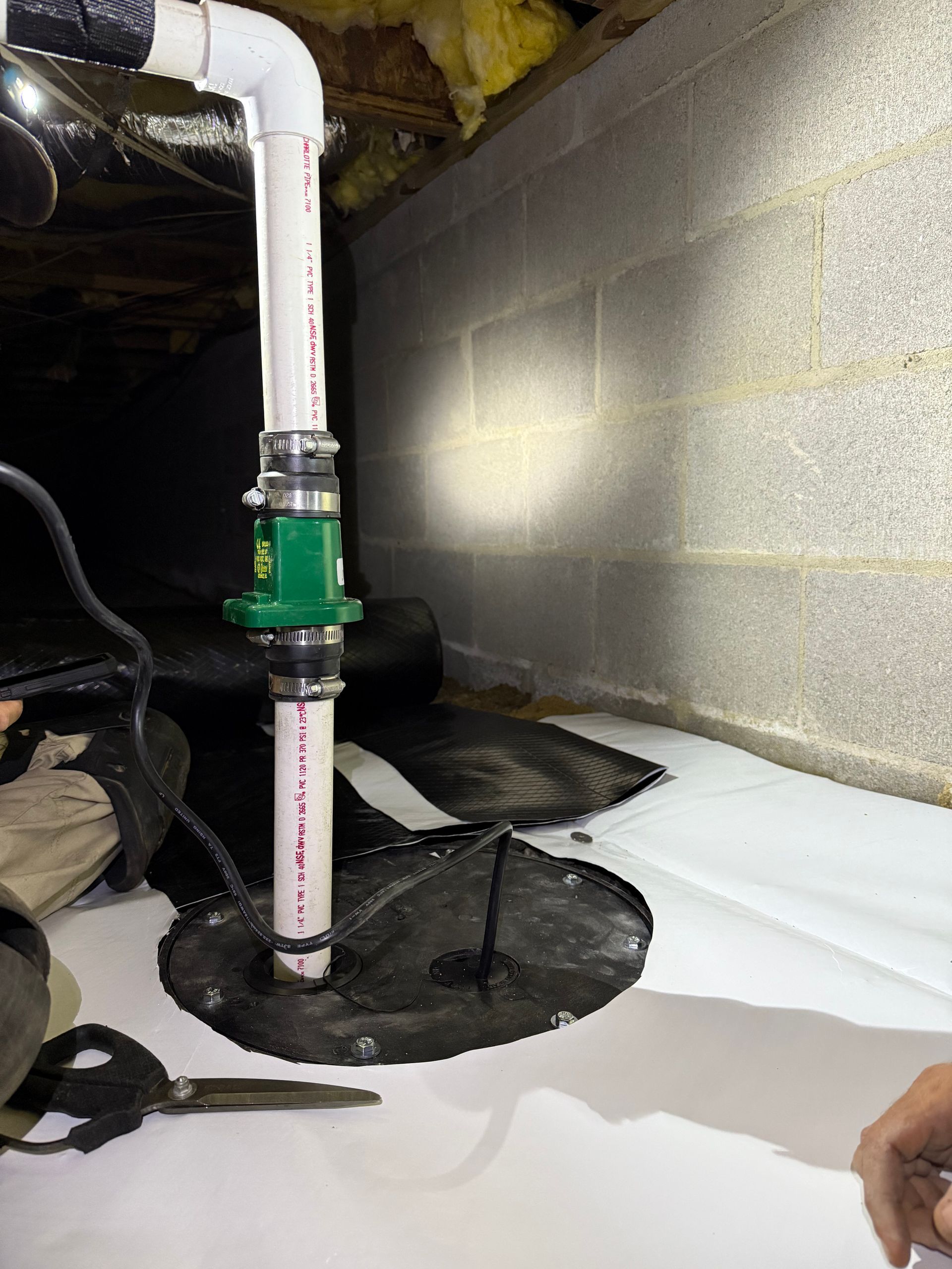 Plumbing pipe assembly with green valve beside a basement wall and floor opening, surrounded by white insulation.