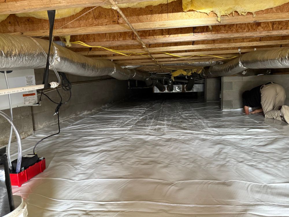 Crawlspace with reflective vapor barrier on floor, exposed insulation overhead, and a worker kneeling at right.