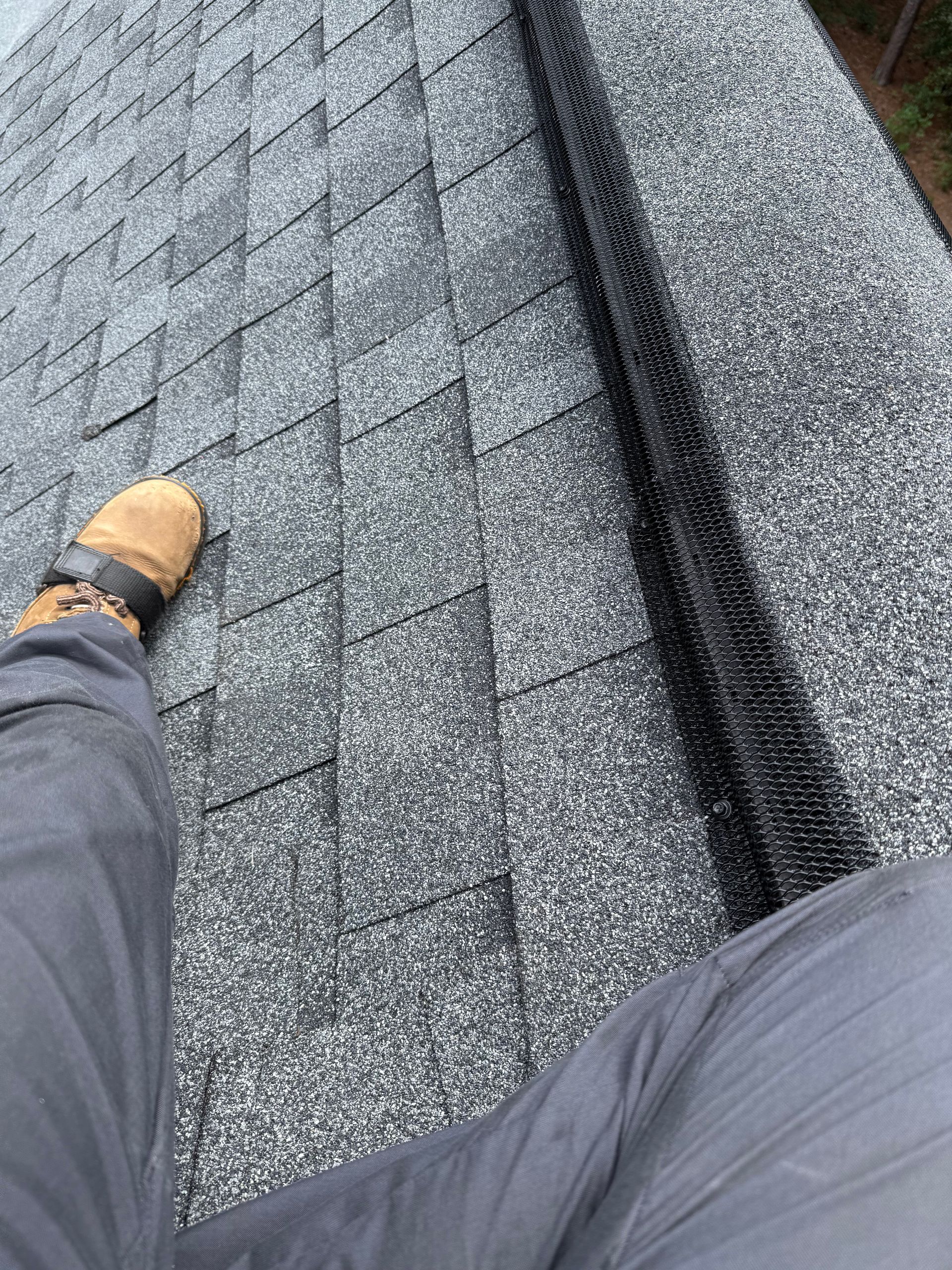 Boot on a shingled roof beside a roof valley, with a person’s legs visible in the foreground
