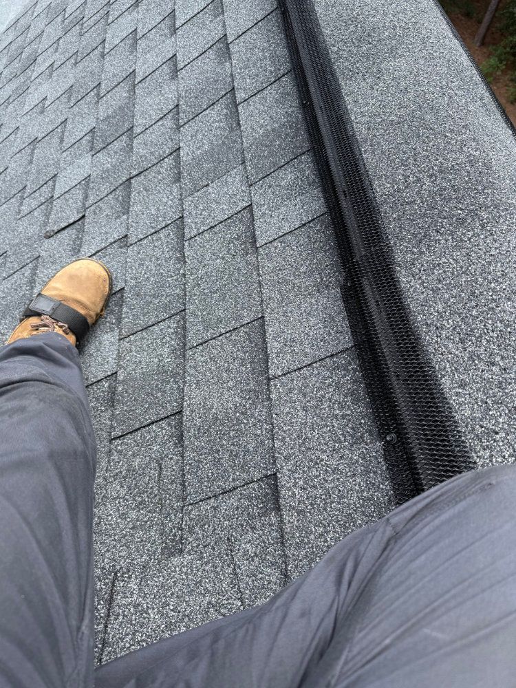 Worker standing on a gray shingled roof beside a dark ridge vent, wearing tan boots and gray pants.