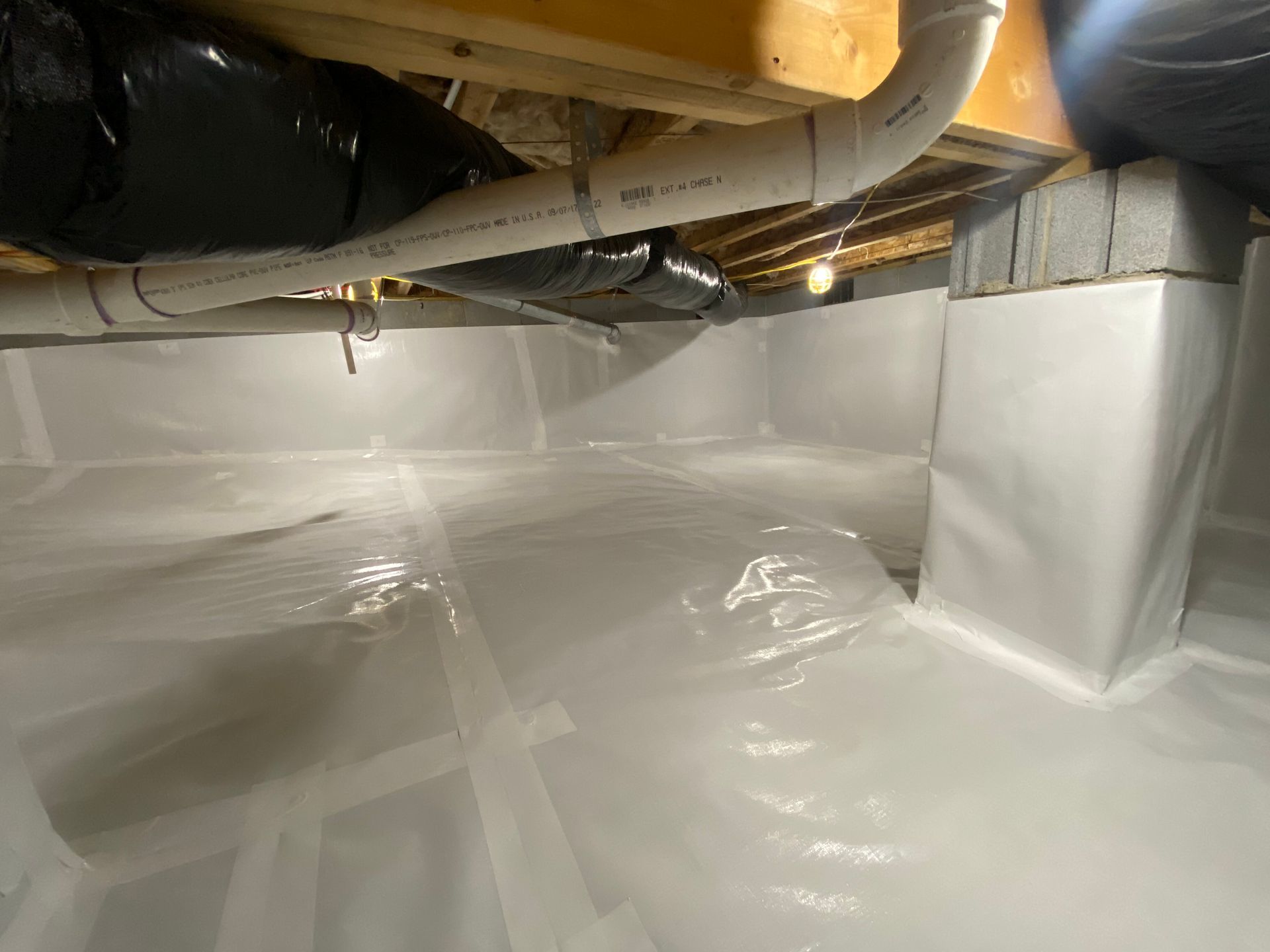 Crawlspace with white vapor barrier lining floor and walls, exposed pipes and ductwork overhead