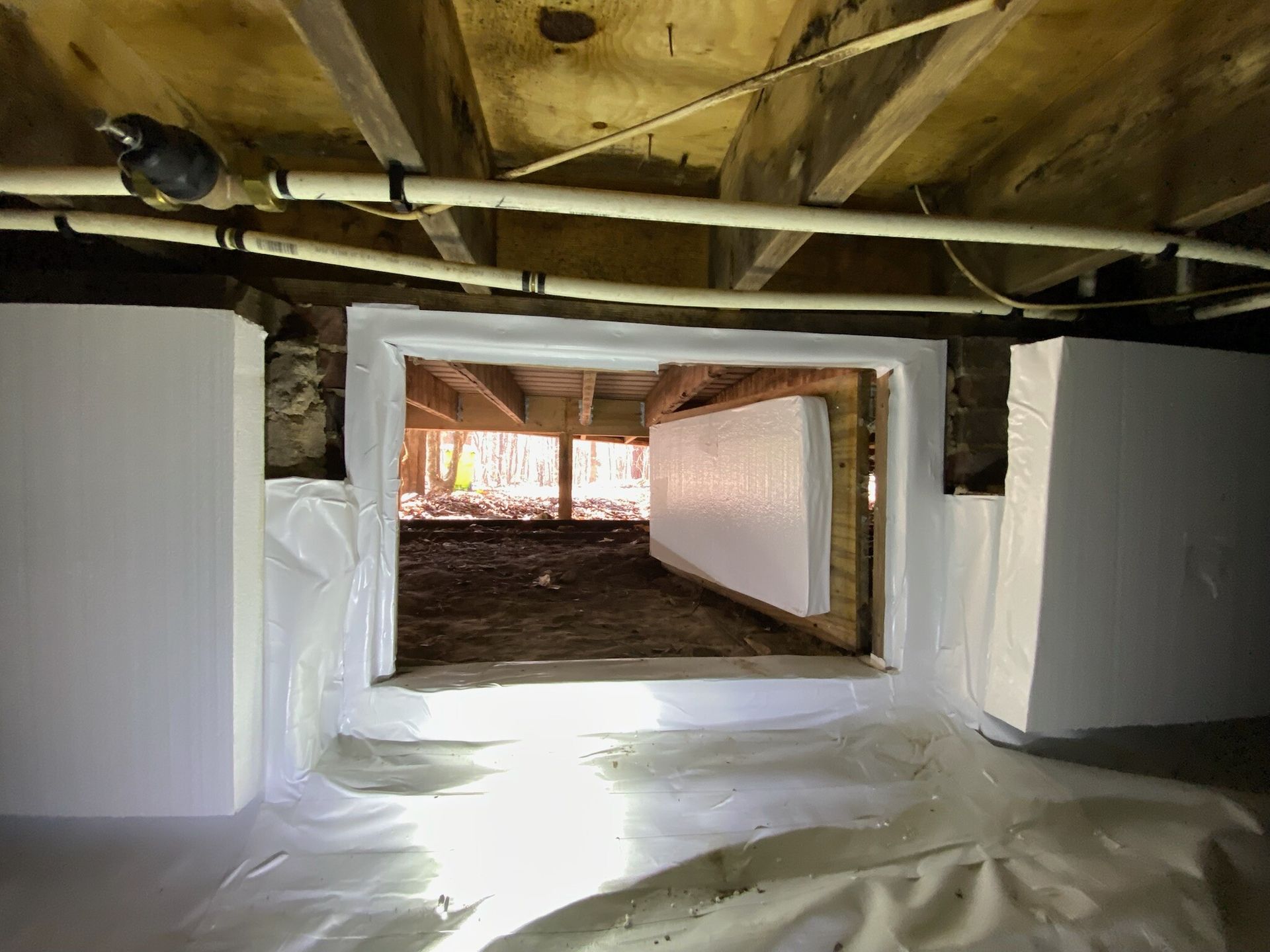 Crawl space with exposed ceiling beams, white insulation, and an opening to the outside.