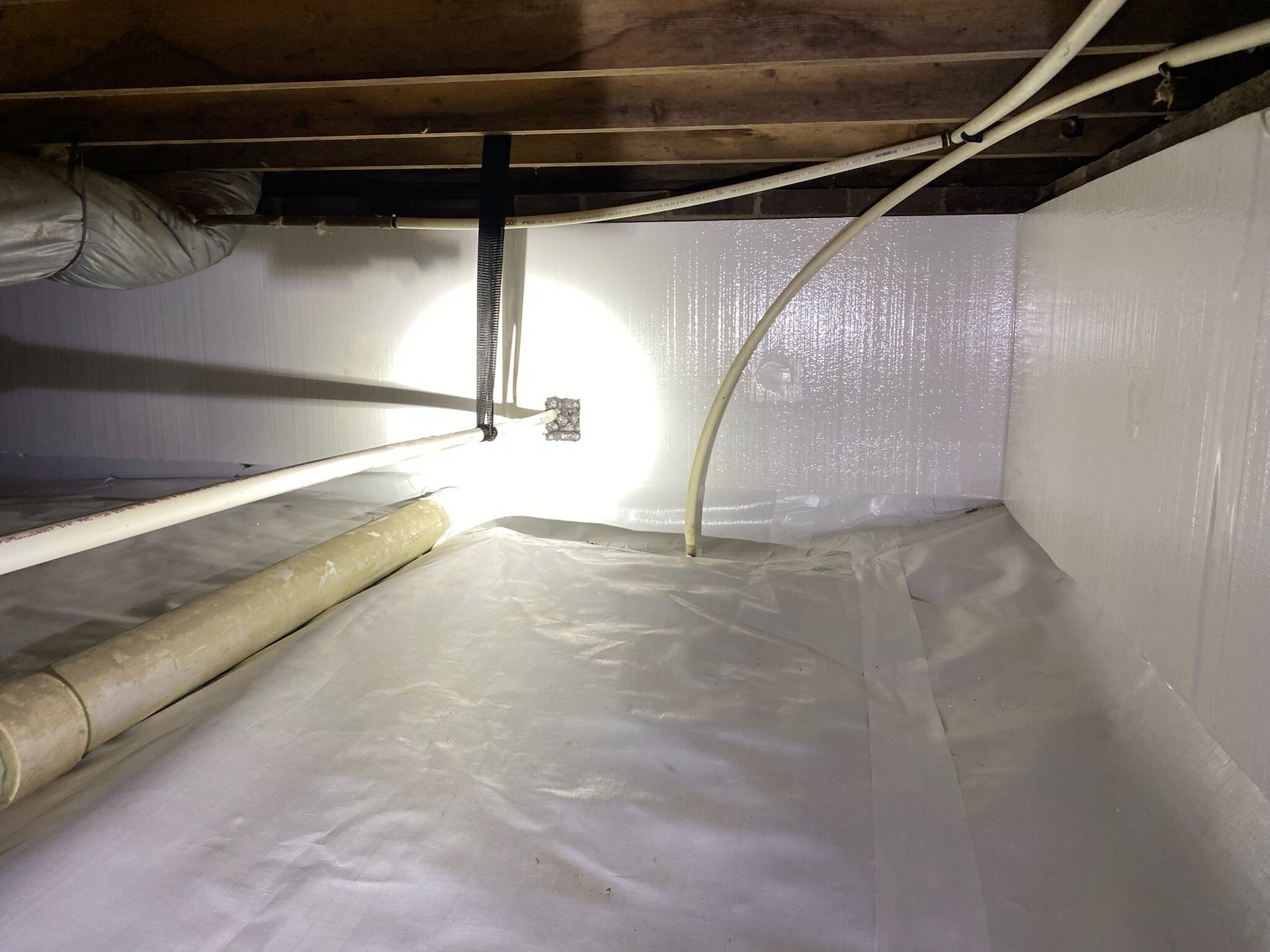 Plastic-lined crawl space with pipes, insulation, and a bright light on the left.