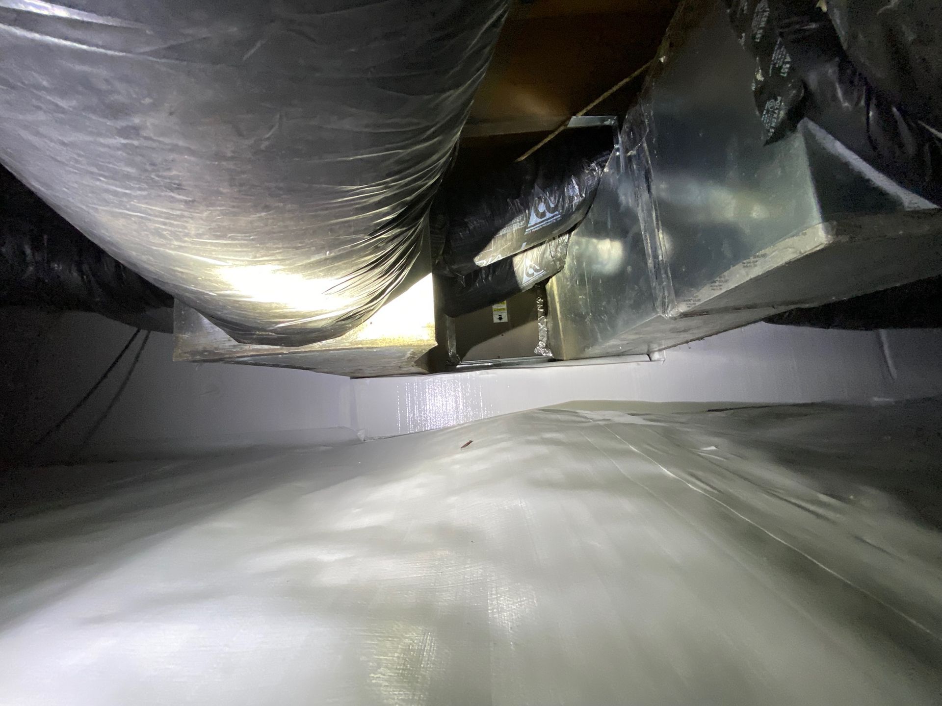Inside an HVAC duct, silver insulated tubes and reflective metal panels in a dim crawlspace