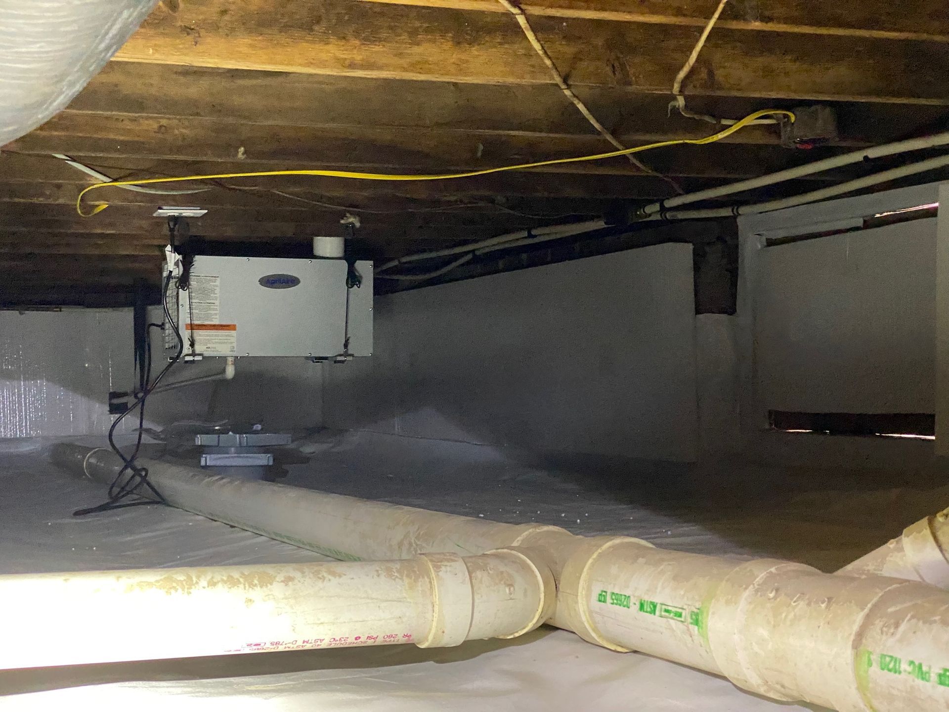 Crawlspace with exposed insulation, pipes, ductwork, and a furnace unit in a dim, narrow area