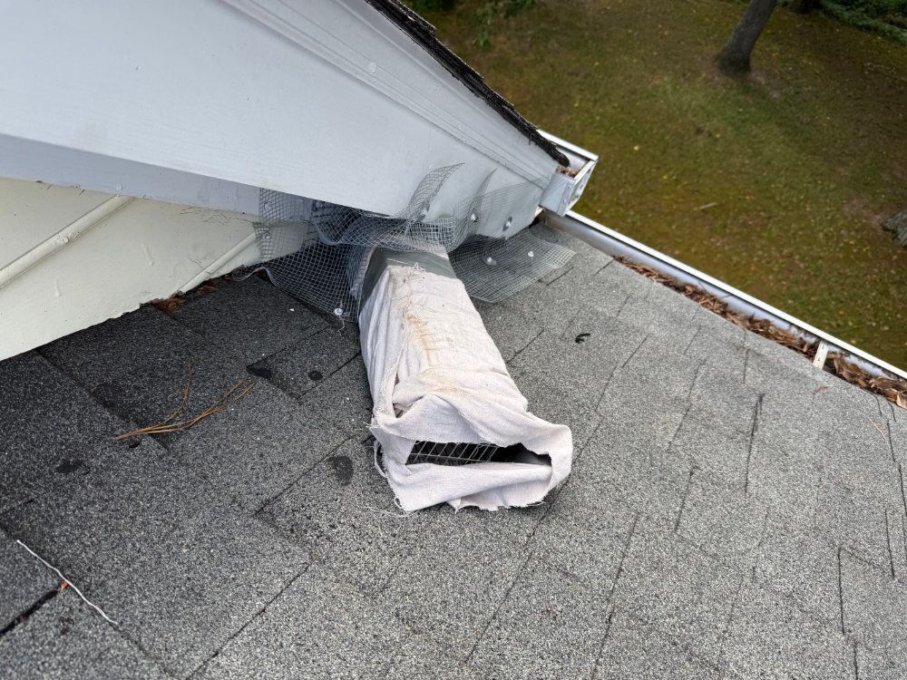 Roof vent pipe wrapped in white insulation on a gray shingled rooftop near the edge of a white roof structure