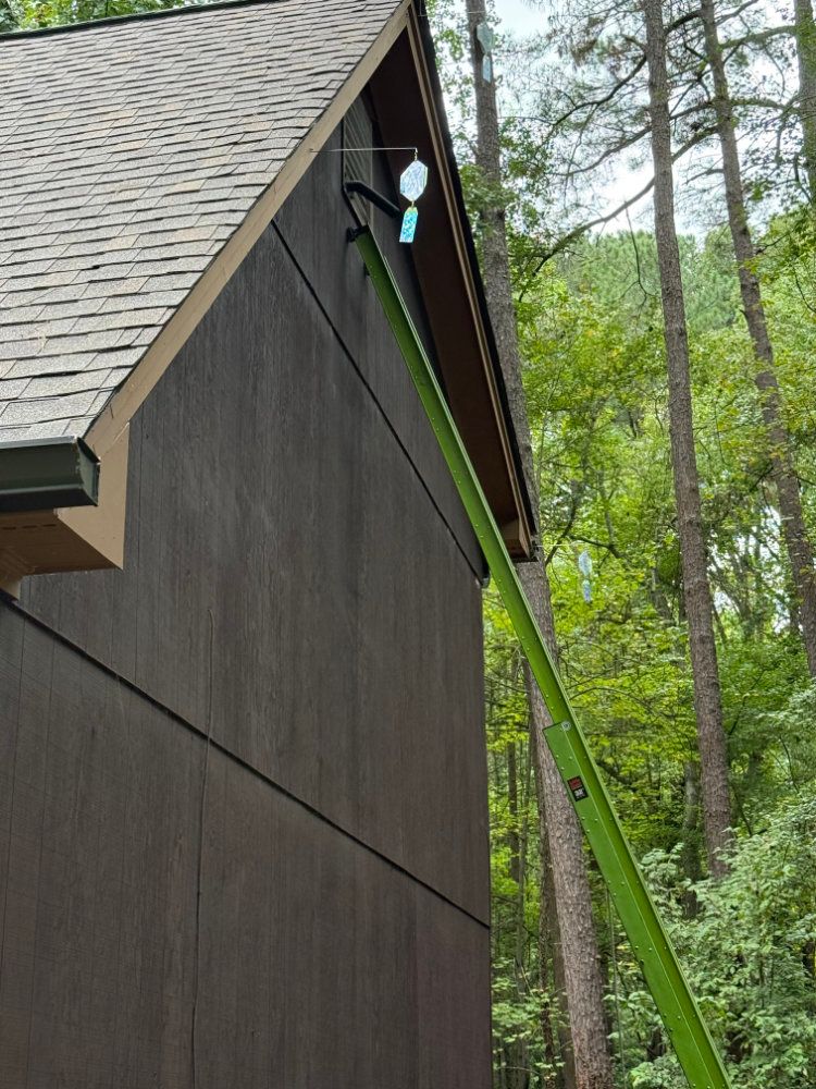 Side of a dark house with a steep shingled roof and green trees in the background