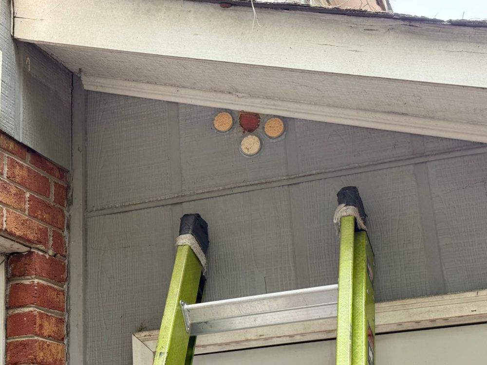 Ladder leaning against house eaves beneath three small circular vents on gray siding