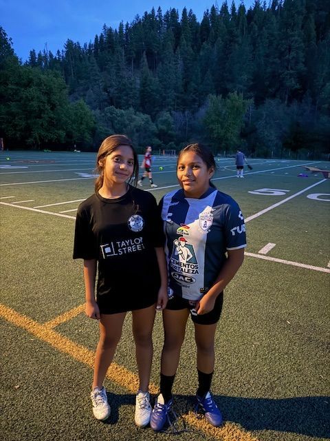 Two young people standing on a sports field. One wears a black shirt, the other a blue and white jersey.