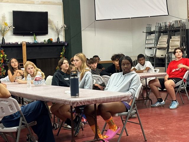 Group of teens sitting at tables, listening to presentation, inside a room.