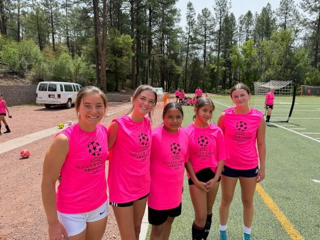 Five young people in pink soccer jerseys stand on a field, smiling.