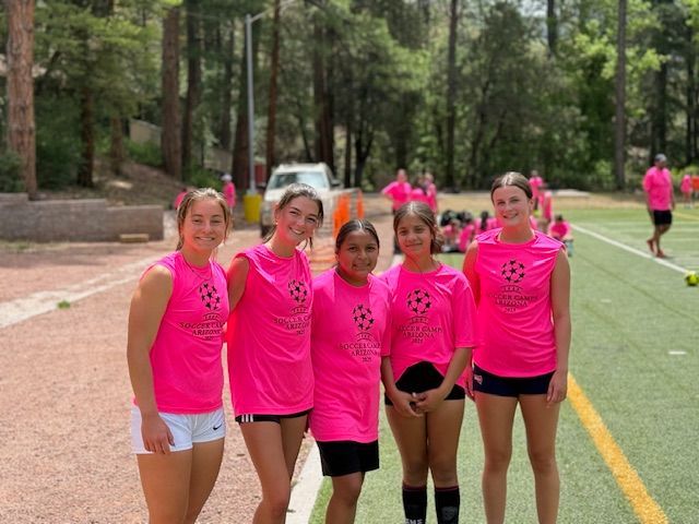Five girls in pink soccer shirts and shorts smile on a field; other people in pink shirts are in the background.