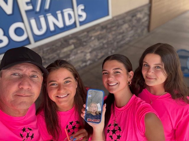 Family of four in pink shirts smiling, holding a phone with a photo in front of a blue sign.