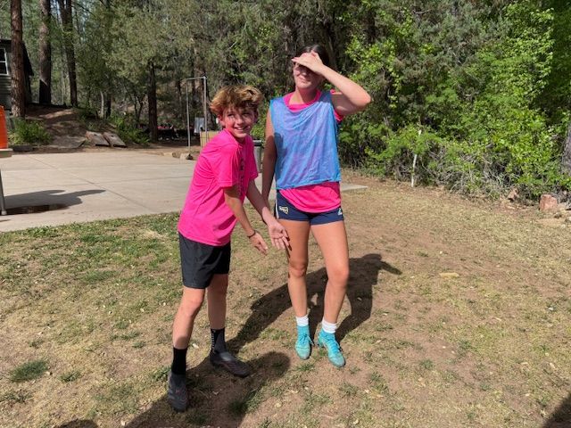 Two people in athletic wear on grass outdoors; one shielding eyes, the other gesturing toward the viewer.