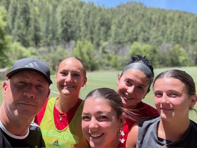Group selfie in front of a green field and trees. Five people smiling at the camera on a sunny day.