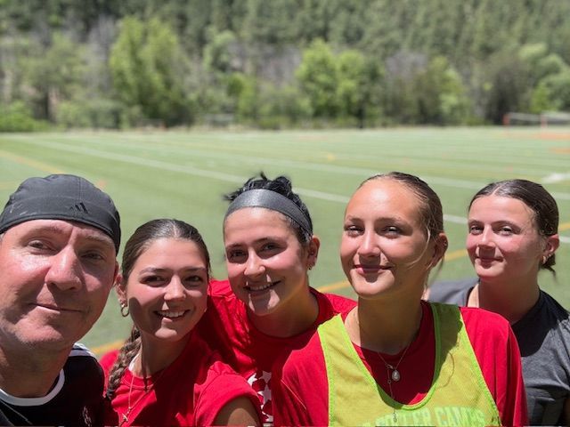 Group of five smiling people, including one with a black cap, pose in front of a green sports field.