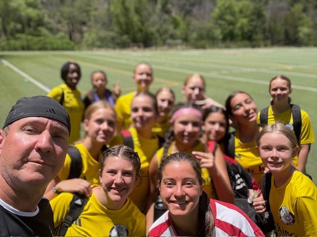 Soccer team in yellow jerseys on a green field pose for a group selfie with two adults.
