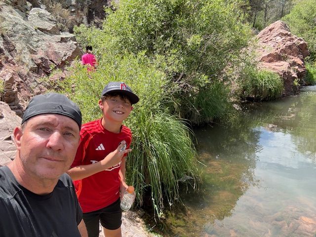 Man and boy smiling next to a stream; rocky background, green foliage.