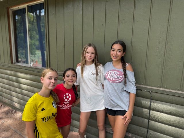 Four girls stand outside a wooden building. One in a yellow shirt, red shirt, white shirt and gray Dr. Pepper shirt.