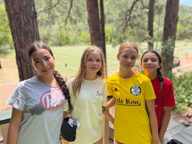 Four girls smiling outdoors near trees; one wears a yellow jersey.