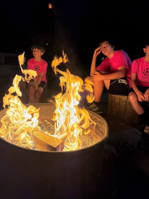 Three people sit by a bonfire at night, flames are bright. All wearing pink shirts.