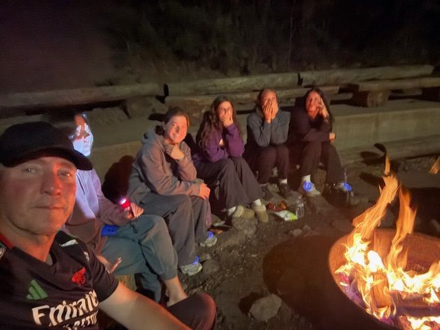 Group of people sitting around a fire at night, some with hands on their faces. Man takes selfie in front.