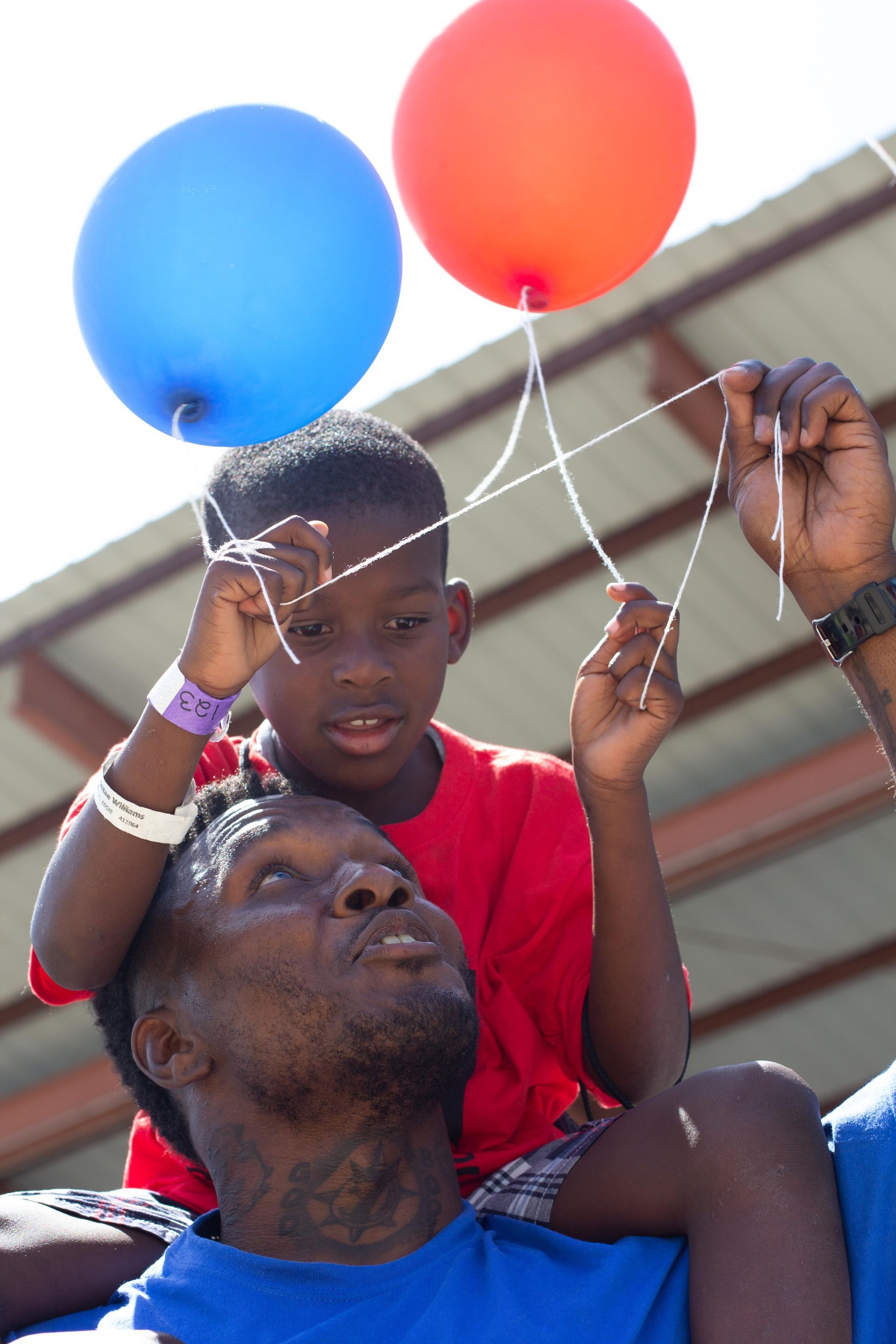 A person carries a child on their shoulders, who holds strings attached to a red and a blue balloon.