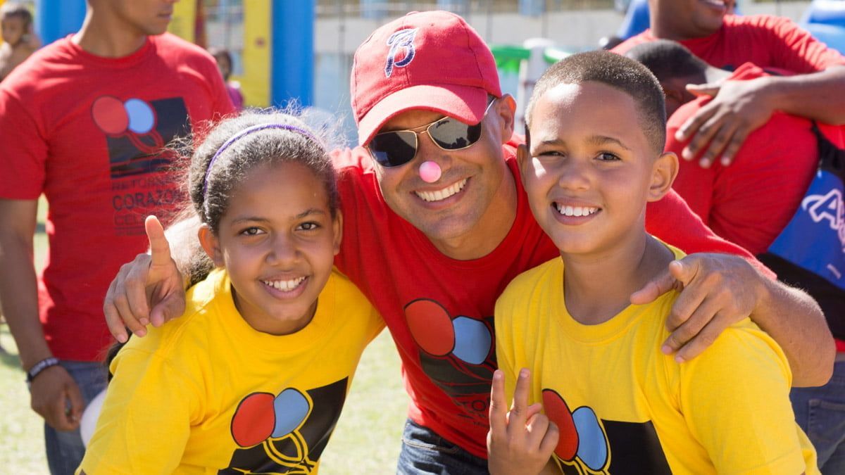 A person wearing a red cap and shirt hugs two smiling children in yellow shirts, all posing outdoors in bright sunlight.