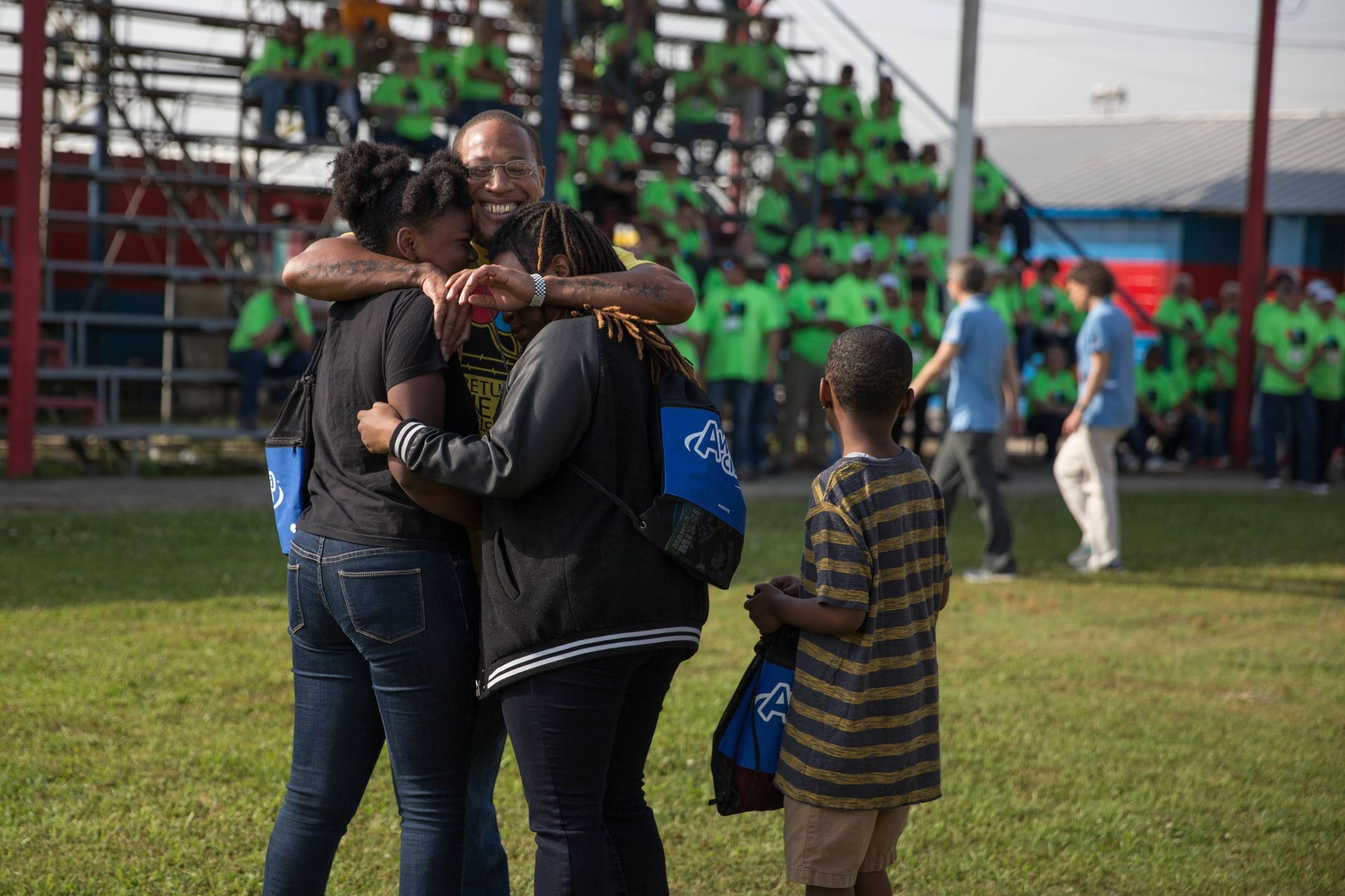 A person stands in a field hugging two others, while a child watches, in front of a crowd in a stadium.
