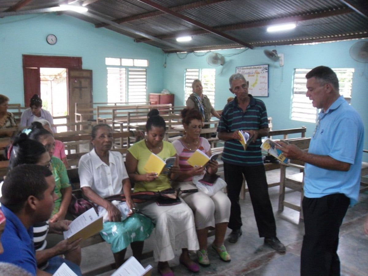 A group of people sitting and standing in a room holding papers while listening to a man speaking.