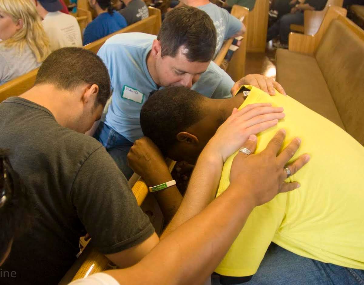 A group of people with their hands on each other’s shoulders, praying together in a church pew.