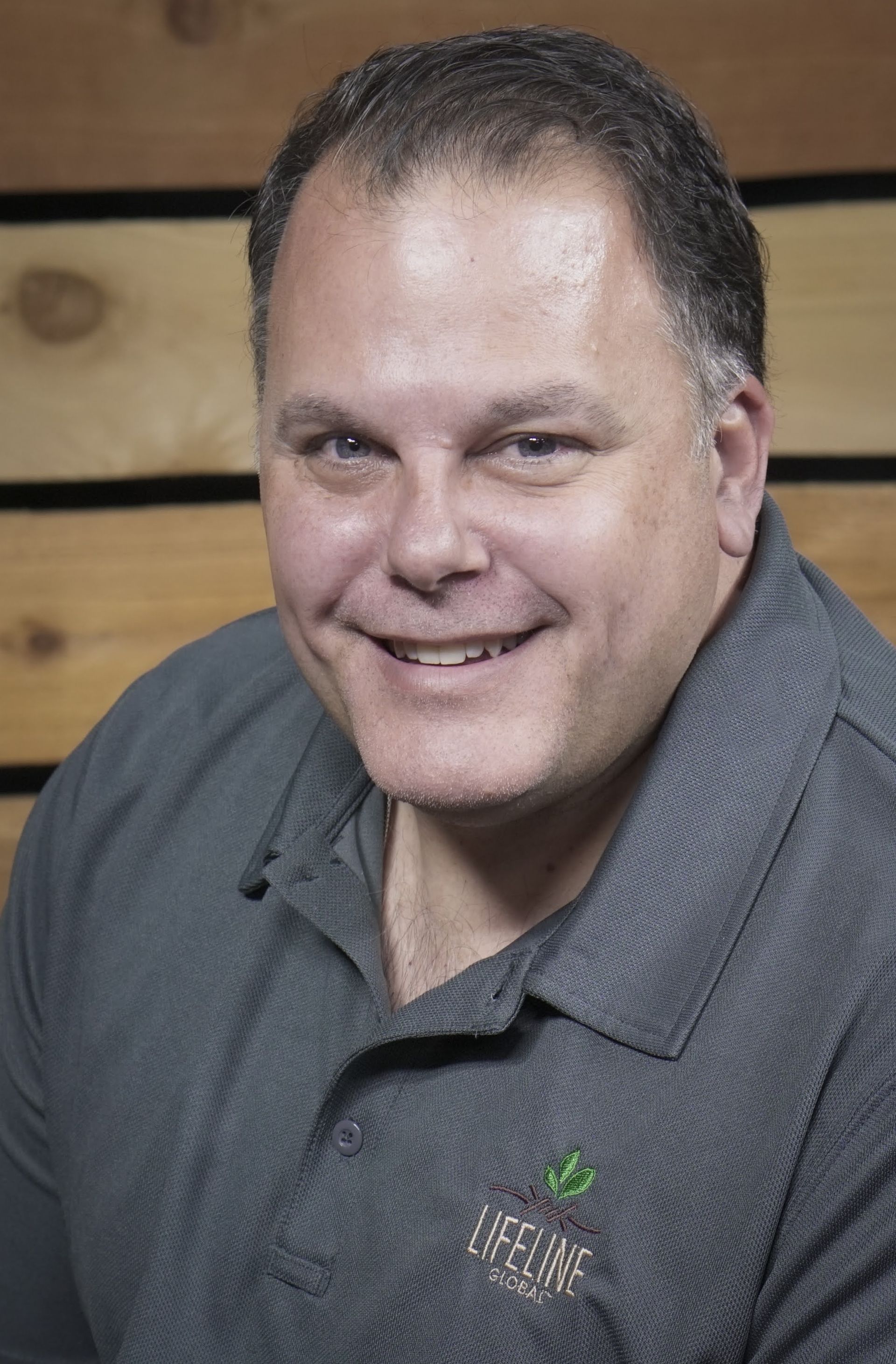 A smiling person wearing a dark grey polo shirt with a logo, positioned against a background of horizontal wood panels.