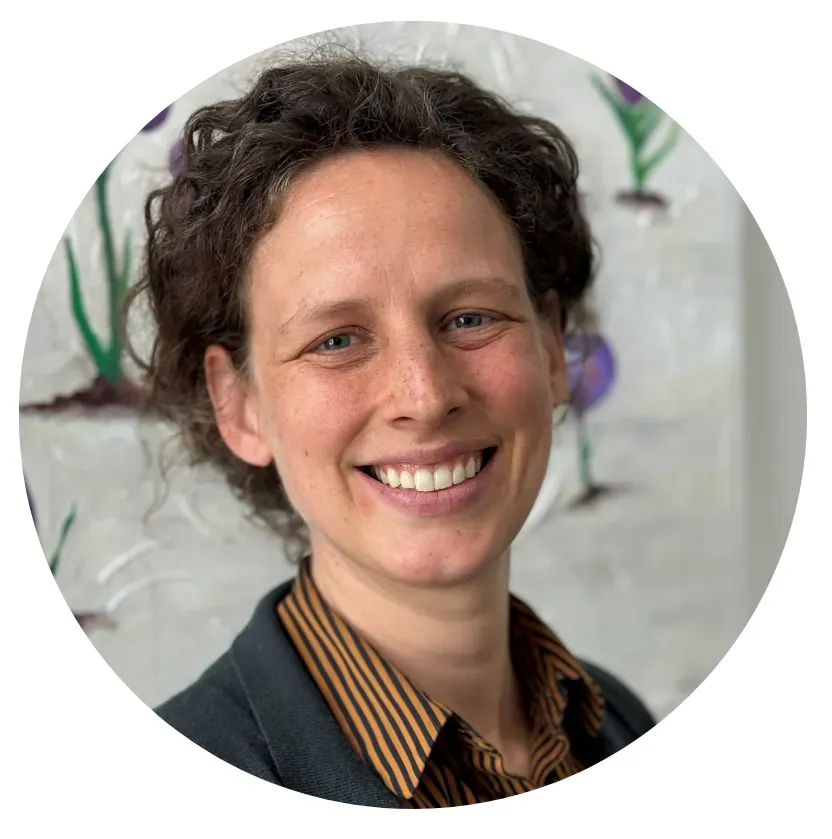 Woman with curly brown hair smiling in front of a painting. Wearing a brown striped shirt and dark blue jacket.