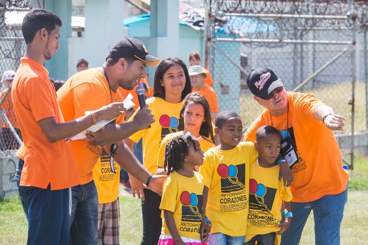 People in orange and yellow shirts participate in an outdoor event, with one person pointing and another using a mic.