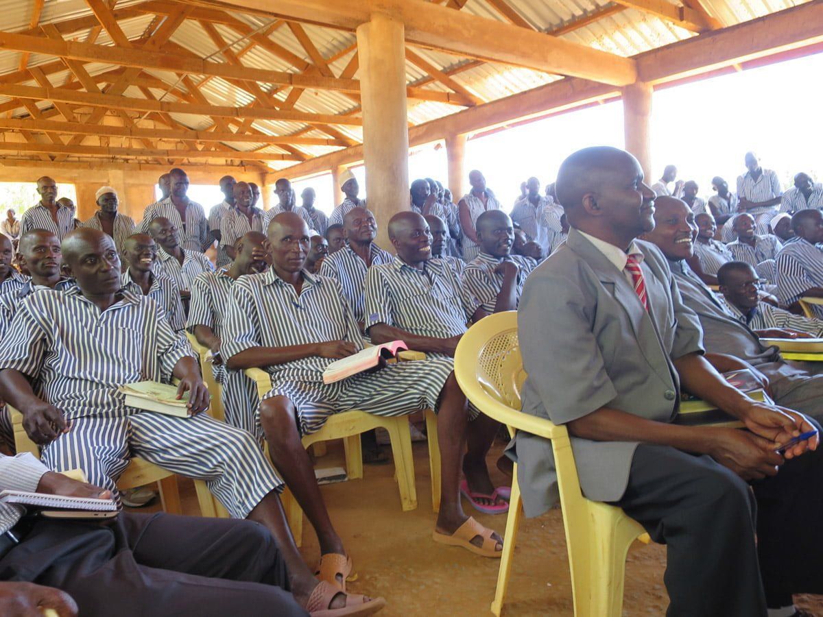 A group of people wearing striped uniforms sit in a large, open-air structure while listening to a speaker.