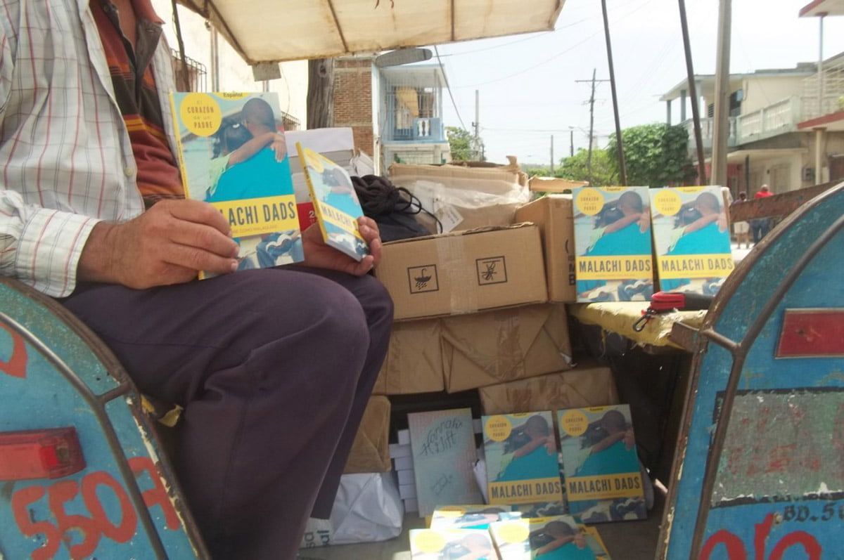 A person sits on a cart selling books, holding copies of a blue-covered title, with more stacked in boxes nearby.