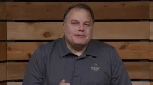 A person in a gray polo shirt stands in front of a horizontal wood-paneled wall, looking toward the camera.