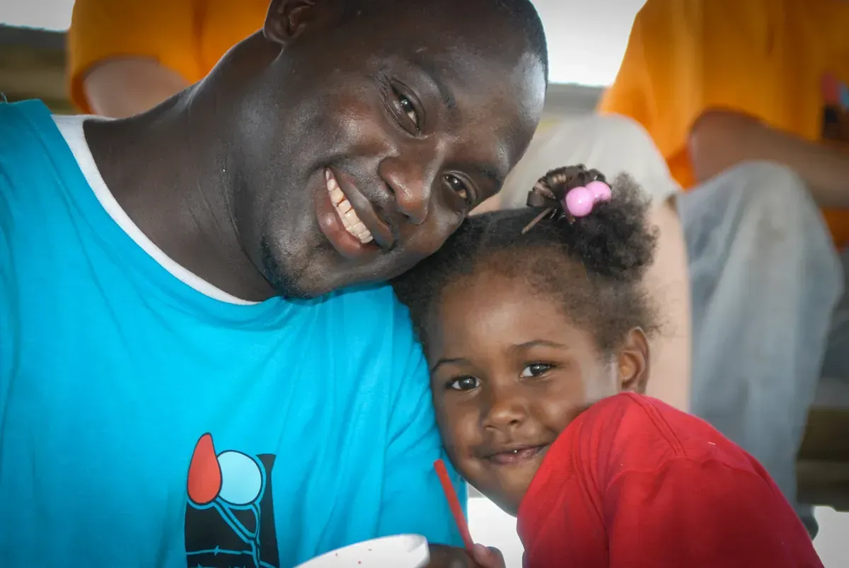 A person in a blue shirt smiling while leaning their head against a child in a red shirt, both looking at the camera.
