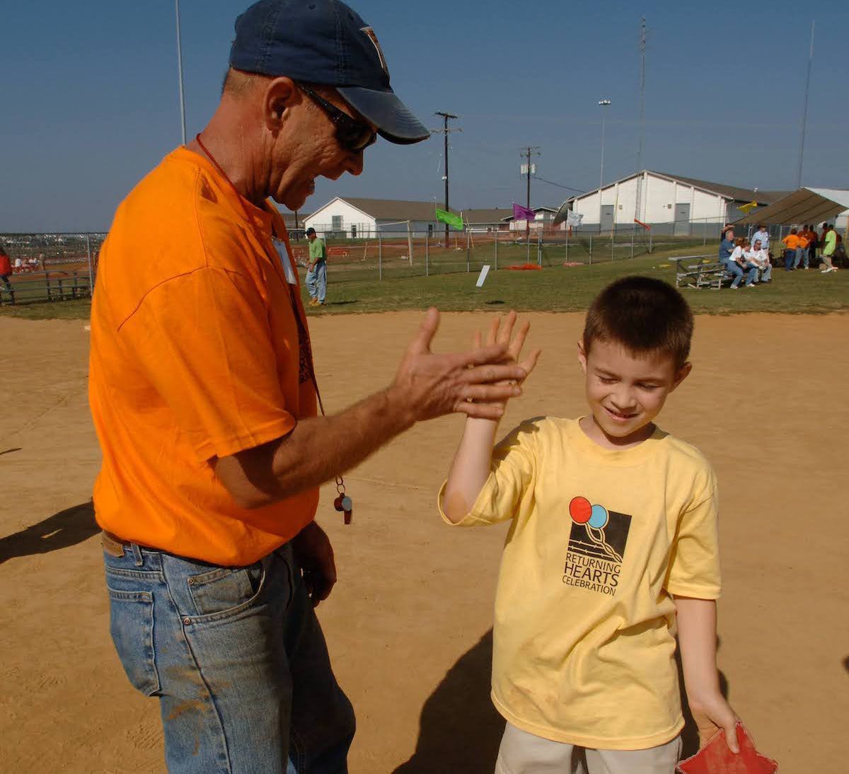 A coach in an orange shirt and a child in a yellow shirt share a high-five on a baseball field.