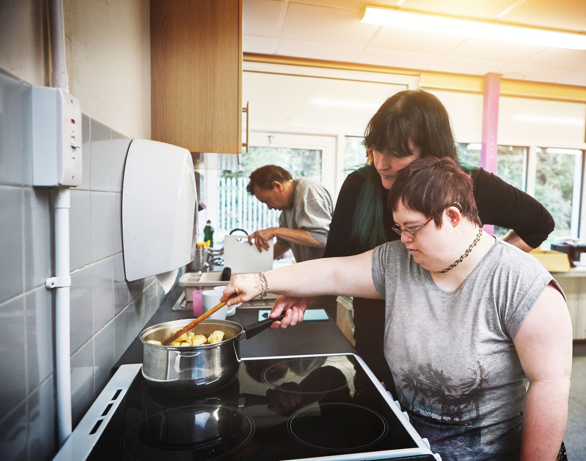 Woman cooking with assistance in a kitchen; another person washes dishes.