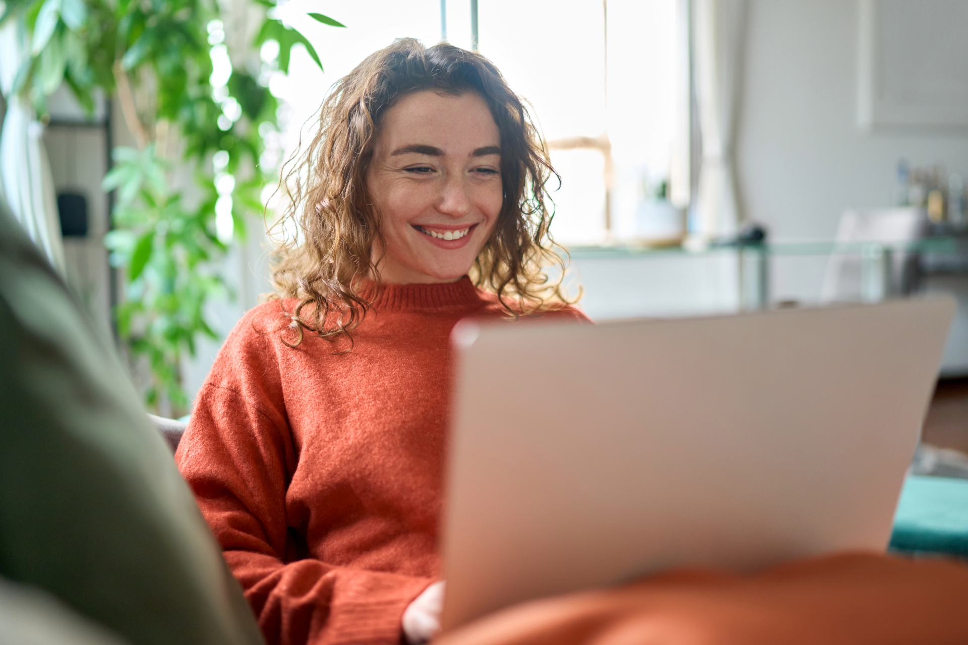 Smiling person in an orange sweater using a laptop indoors by a bright window