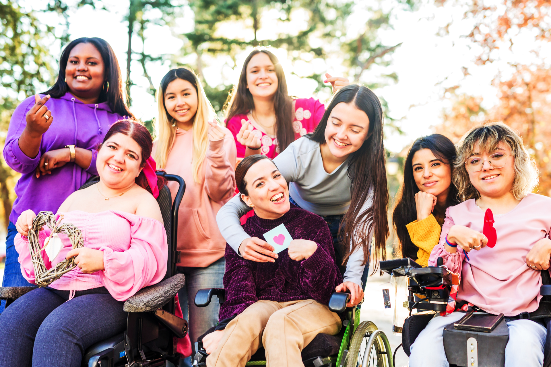 Group of smiling people in wheelchairs holding flowers outdoors in a park, with autumn trees in the background