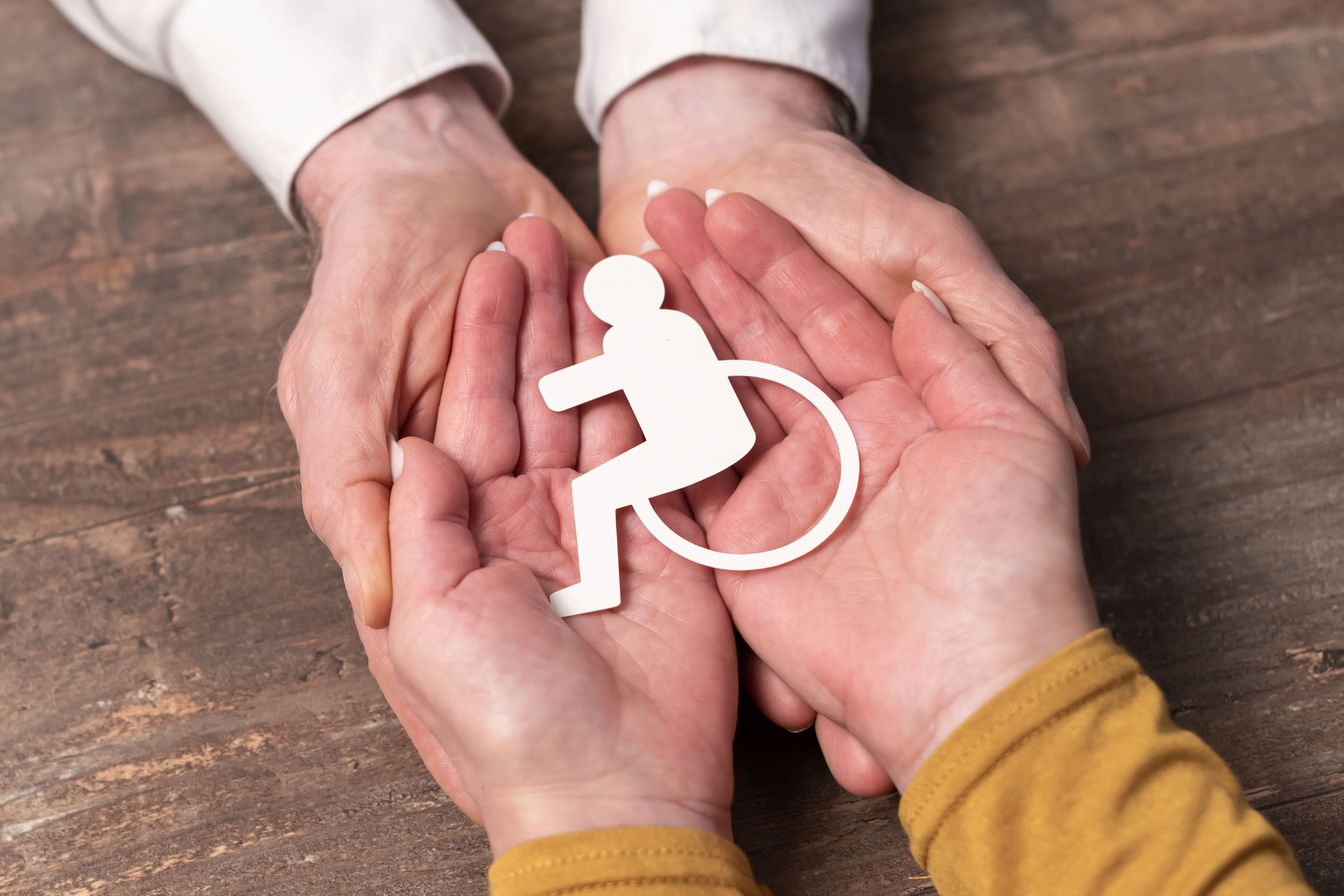 Hands cupping a white wheelchair access symbol on a wooden table