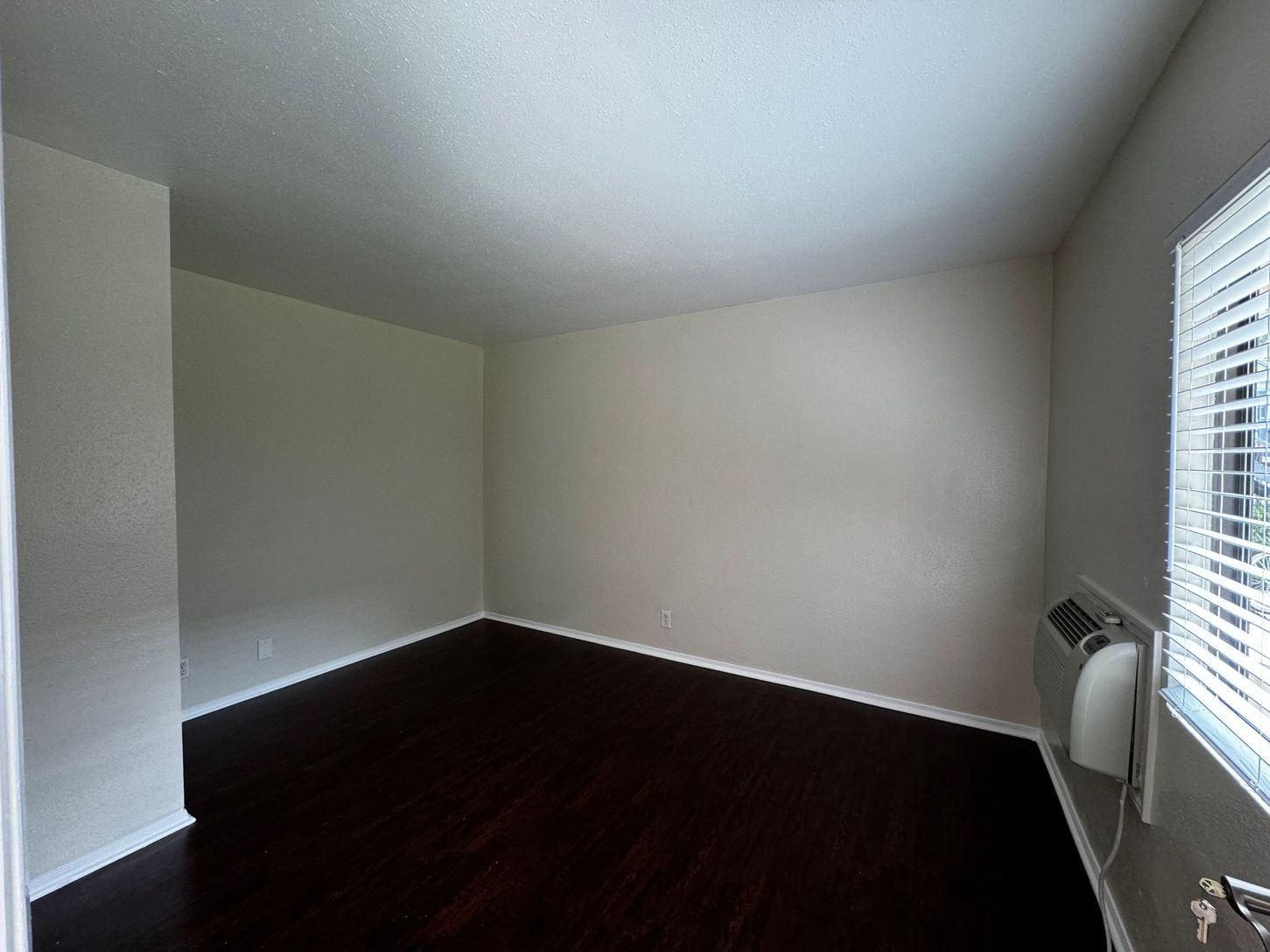 An empty living room with hardwood floors and a window with blinds.