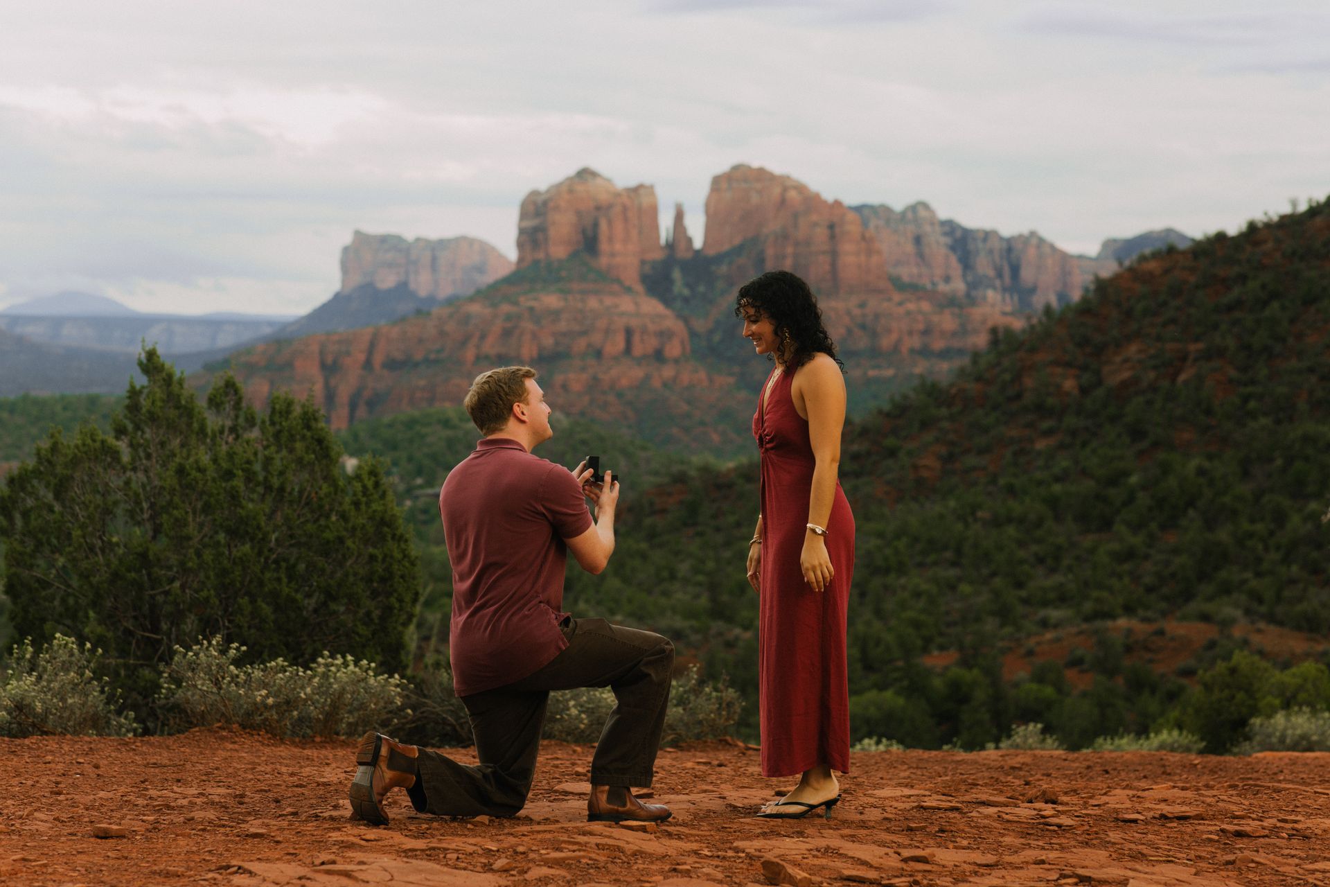 Two people on a rocky ledge before red sandstone cliffs under an overcast sky