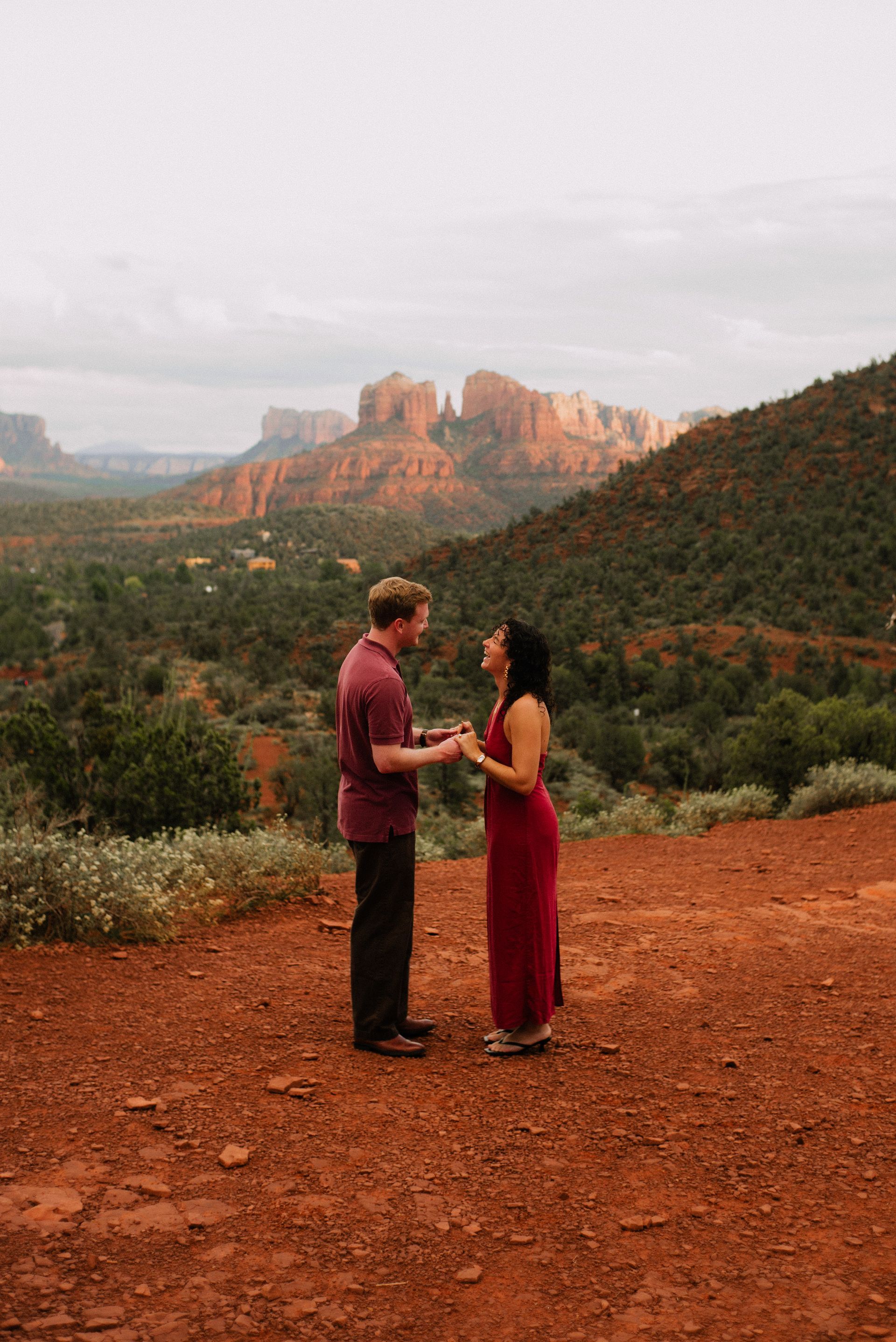 Person holding a small dog in front of red rock cliffs at a desert overlook