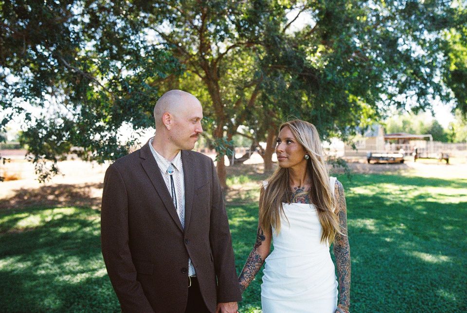 Couple walking hand in hand in a sunny park, with trees and grass in the background.